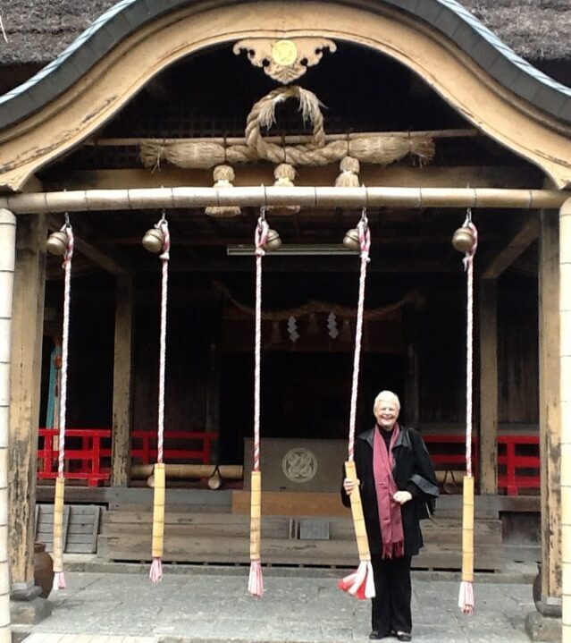 Linden Thorp writer and strategist is a blond haired, formally-dressed woman standing holding a decorated bell-rope outside a temple entrance in Japan